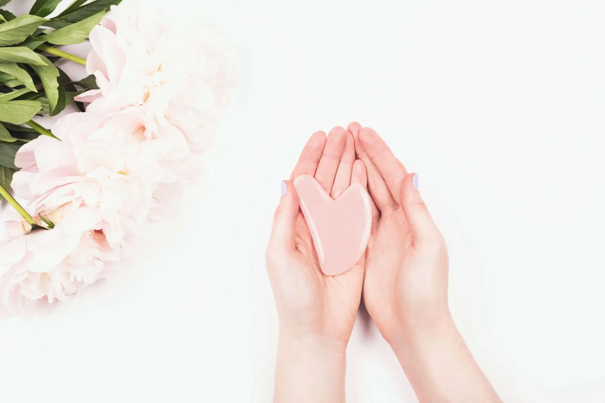 Valerie Millet Dos manos sosteniendo un objeto rosa en forma de corazón sobre un fondo blanco, con flores de color rosa claro en el lado izquierdo. Bienestar Holistico