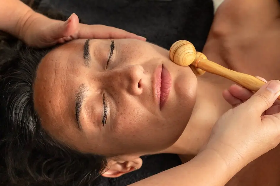 Valerie Millet Una persona con los ojos cerrados recibe un masaje facial con una herramienta de madera, que encarna el bienestar global, mientras alguien le sostiene suavemente la cabeza. Bienestar Holistico