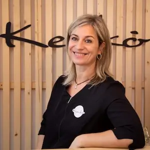 Valerie Millet Una mujer de cabello rubio sonríe, parada frente a un cartel con la palabra "Keiko" de fondo. Lleva una blusa negra y una insignia con un círculo y un texto, que recuerda el estilo único de Valerie y ofrece un testimonio auténtico a través de su expresión. Bienestar Holistico