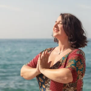 Valerie Millet Una mujer con cabello castaño corto y un vestido colorido está parada junto al mar con las manos en posición de oración, los ojos cerrados, sonriendo bajo un cielo despejado, encarnando el bienestar global. Bienestar Holistico