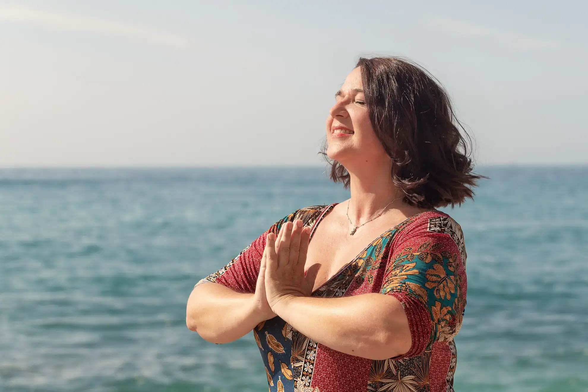 Valerie Millet Una mujer con cabello castaño corto y un vestido colorido está parada junto al mar con las manos en posición de oración, los ojos cerrados, sonriendo bajo un cielo despejado, encarnando el bienestar global. Bienestar Holistico