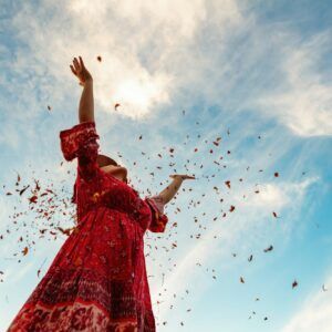 Valerie Millet Una persona con un vestido rojo está de pie con los brazos en alto mientras las hojas caen a su alrededor contra un cielo azul con nubes. Bienestar Holistico