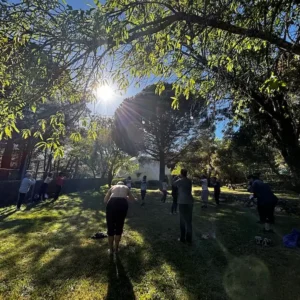 Valerie Millet Personas que participan en una sesión de ejercicio al aire libre en una zona de césped bajo la luz del sol, rodeada de árboles, similar a retiros de empoderamiento. Bienestar Holistico