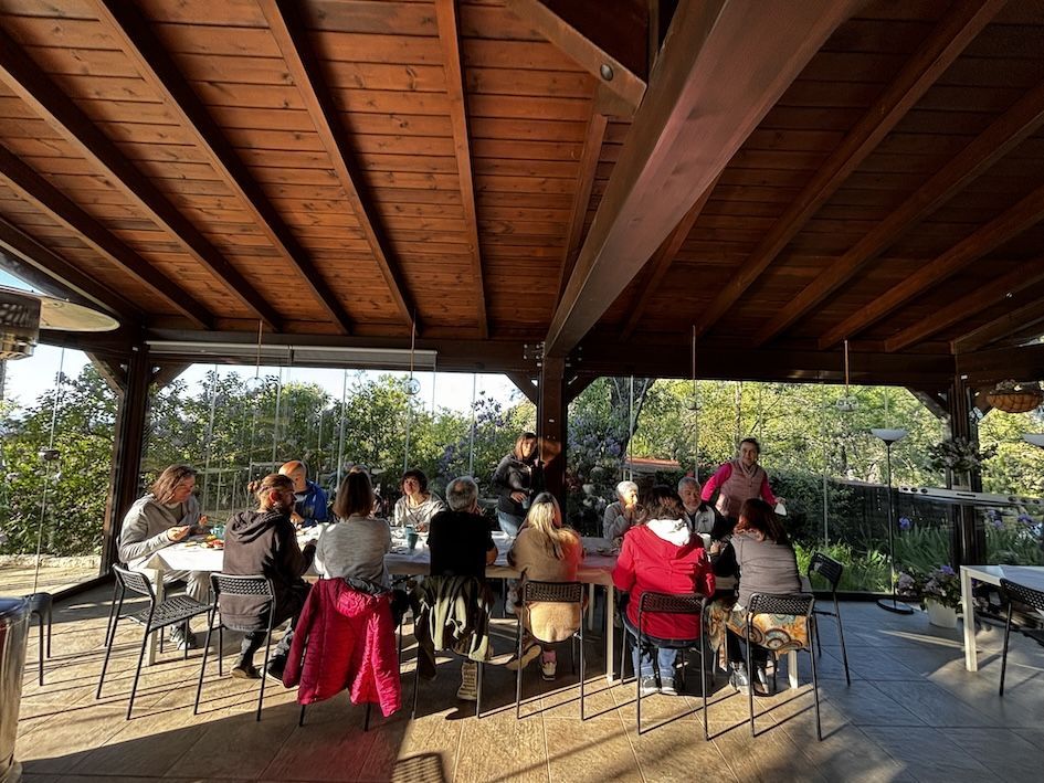 Valerie Millet Un grupo de personas sentadas en una mesa larga bajo un pabellón de madera en un ambiente al aire libre, conversando y cenando, disfrutando de la camaradería que a menudo se encuentra durante los retiros de empoderamiento. Bienestar Holistico