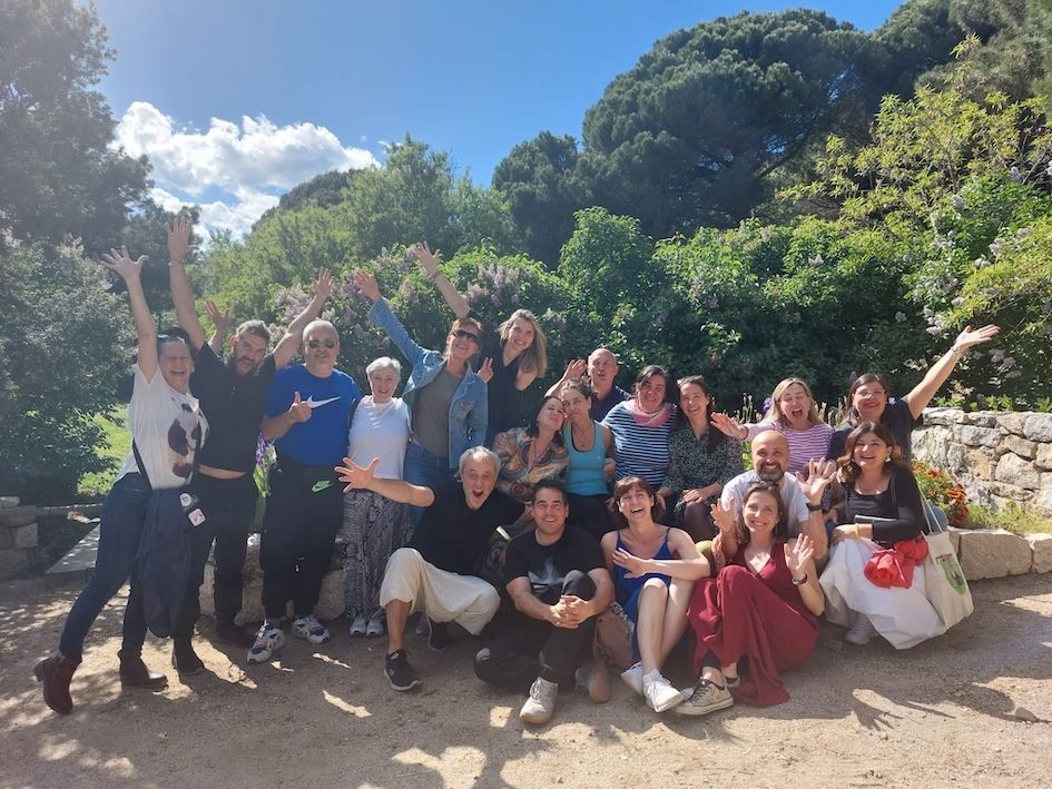 Valerie Millet Un grupo de personas paradas y sentadas al aire libre con árboles y arbustos al fondo, todos sonriendo y levantando las manos con alegría. Bienestar Holistico
