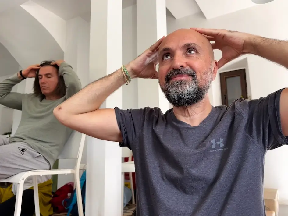 Valerie Millet Dos personas están sentadas en el interior con los ojos cerrados, las manos sobre la cabeza y parecen estar en un estado meditativo o relajado. El fondo presenta paredes blancas y elementos de diseño arqueados, que recuerdan a los espacios utilizados para los retiros de empoderamiento. Bienestar Holistico
