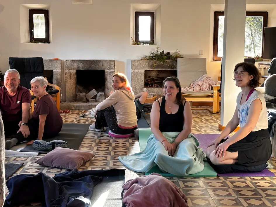 Valerie Millet Un grupo de personas sentadas sobre colchonetas de yoga en una habitación bien iluminada con un suelo estampado y ventanas al fondo. Algunos sonríen y a la izquierda se ve una chimenea. Bienestar Holistico