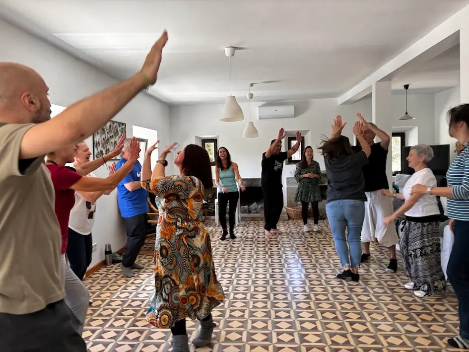 Valerie Millet Un grupo de personas se paran en círculo con los brazos en alto, participando en una actividad animada dentro de una habitación con suelos de baldosas estampadas y paredes blancas. Bienestar Holistico