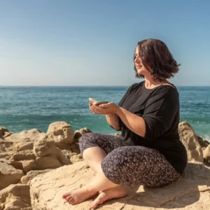 Valerie Millet Una mujer sentada con las piernas cruzadas en una orilla rocosa junto al océano, sosteniendo un objeto en sus manos, con el mar y el cielo despejado de fondo, capturando un momento sereno de paz que a menudo se encuentra en los retiros de empoderamiento. Bienestar Holistico
