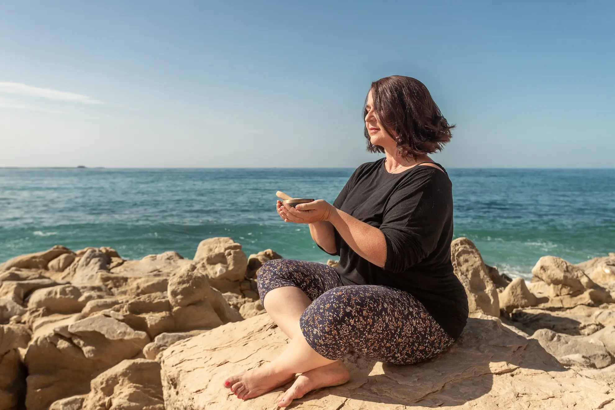 Valerie Millet Una mujer sentada con las piernas cruzadas en una orilla rocosa junto al océano, sosteniendo un objeto en sus manos, con el mar y el cielo despejado de fondo, capturando un momento sereno de paz que a menudo se encuentra en los retiros de empoderamiento. Bienestar Holistico