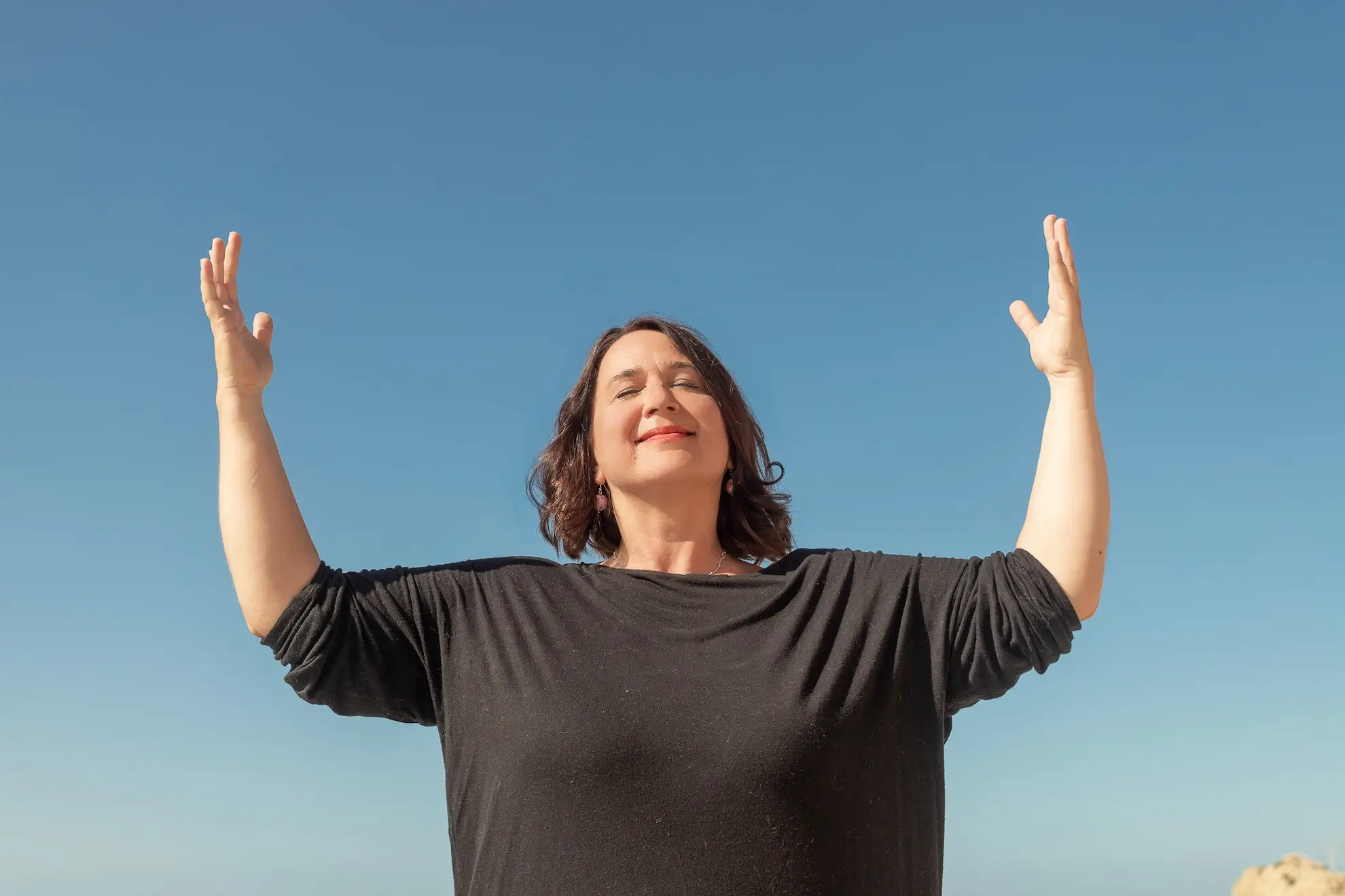 Valerie Millet Una persona con los ojos cerrados y los brazos extendidos se alza contra un cielo azul claro. Bienestar Holistico