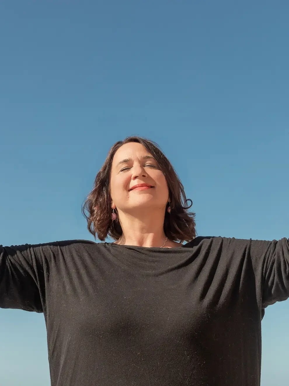 Valerie Millet Una persona con los ojos cerrados y los brazos extendidos se alza contra un cielo azul claro. Bienestar Holistico