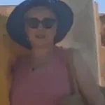 Valerie Millet Una persona con gafas de sol y un sombrero de ala ancha se encuentra al aire libre, sosteniendo una bolsa al hombro. Están vestidos con una blusa sin mangas y parecen estar en un lugar iluminado por el sol, muy parecido a lo que Valerie describió en su testimonio sobre las aventuras de verano. Bienestar Holistico