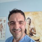 Valerie Millet Un hombre vestido con una bata azul sonríe a la cámara con un historial médico y una pintura de Buda al fondo. Bienestar Holistico